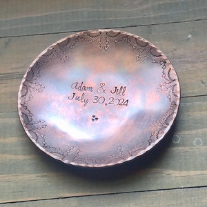 May include: A copper ring dish with a heart design border. The dish is engraved with the text "Adam & Jill July 30, 2024".