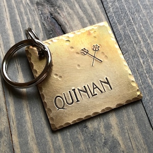 May include: A brass key chain tag with a hammered finish. The tag has a pair of crossed tridents and the name "QUINLAN" engraved on it.
