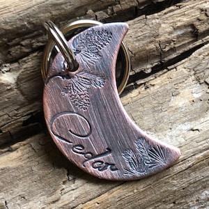 May include: A copper-toned crescent moon-shaped dog tag with a key ring. The tag is engraved with the name "Cedar" and a pine tree design.