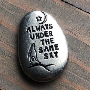 May include: Oval, silver-colored stone with the words "ALWAYS UNDER THE SAME SKY" engraved. A crescent moon and star are above the text, and a rabbit silhouette is below. The stone is on a wooden surface.