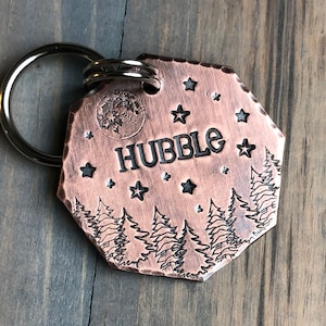 May include: A copper-colored dog tag with a black engraved design of a forest scene with stars and a moon. The tag reads "HUBBLE" in black lettering.