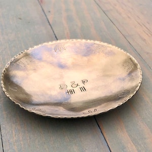 May include: A brass tray with a hammered finish and a scalloped edge. The tray has a decorative design around the edge and the initials "B&P" and "HHH III" are engraved in the center.