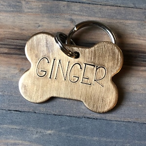 May include: A gold-coloured dog tag shaped like a bone, with the name "GINGER" engraved on it. A silver-coloured split ring is attached for collar attachment. The background is a weathered wooden surface, with a greyish-blue hue.