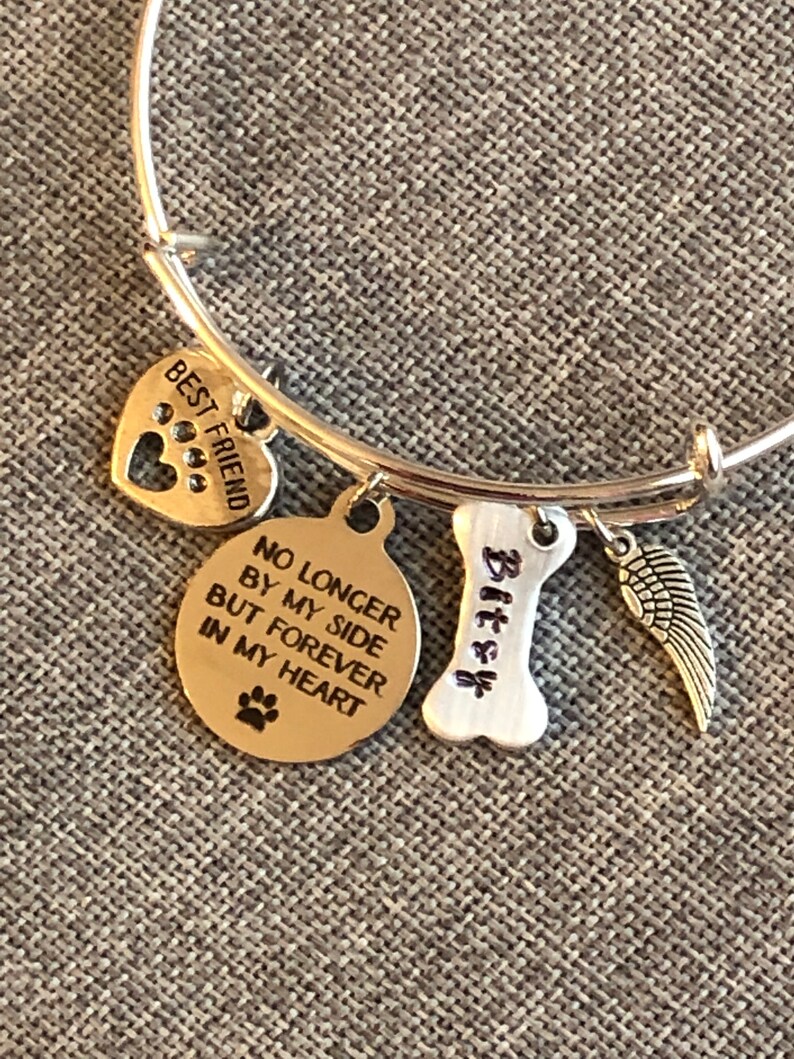 Op de afbeelding: Zilveren armband met bedels die "Beste vriend", "Niet langer aan mijn zijde, maar voor altijd in mijn hart", "Bitzy" en een vleugelbedel zeggen.