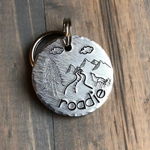 May include: Silver dog tag with a mountain scene and the word "roadie" engraved on it.