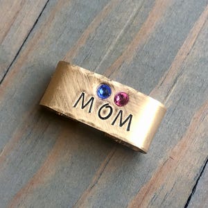 May include: Gold metal charm with the word "MOM" engraved on it. The charm has two small gemstones, one blue and one pink.