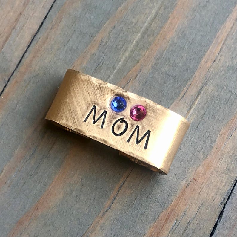 May include: Gold metal charm with the word "MOM" engraved on it. The charm has two small gemstones, one blue and one pink.
