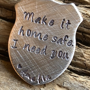 Puede incluir: Un amuleto de metal plateado en forma de escudo con el texto "Make it home safe I need you" y un coraz&oacute;n con el nombre "Amelia" grabado en &eacute;l.