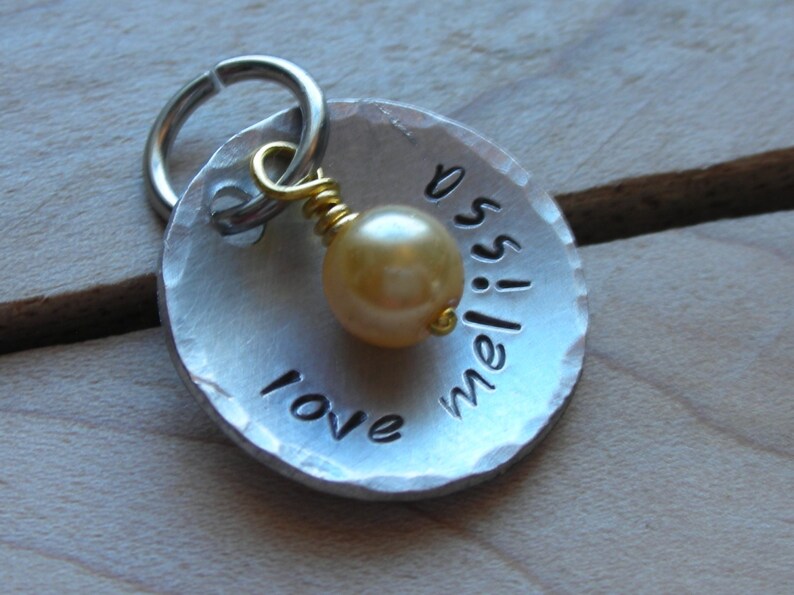 May include: A silver metal dog tag with a hammered finish and a small gold bead. The tag is engraved with the words "love meliss" and has a silver ring for attaching to a collar.