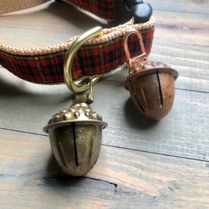 May include: A dog collar with a red and black plaid pattern. The collar has a brass ring and two acorn-shaped bells. One bell is brass-colored, and the other is a rusty brown. The collar is made of a beige woven material.