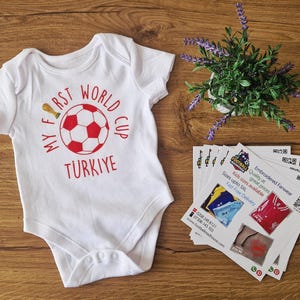 Könnte beinhalten: Weißer Baby-Body mit dem roten Aufdruck "MY FIRST WORLD CUP TÜRKİYE". Das Design zeigt einen Fußball und eine Trophäe. Eine kleine Pflanze und mehrere Karten sind ebenfalls sichtbar.
