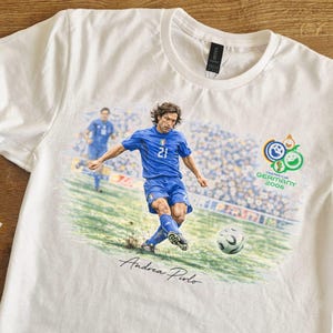 May include: White t-shirt featuring a soccer player in a blue uniform kicking a soccer ball. The shirt has a graphic of the 2006 FIFA World Cup logo and the name "Andrea Pirlo" in script.