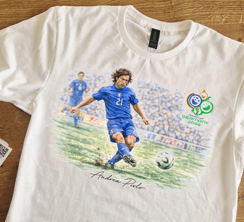 May include: White t-shirt featuring a soccer player in a blue uniform kicking a soccer ball. The shirt has a graphic of the 2006 FIFA World Cup logo and the name "Andrea Pirlo" in script.