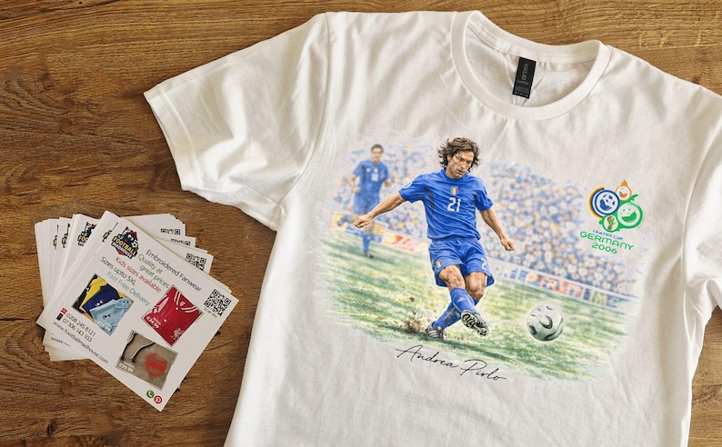 May include: White t-shirt featuring a soccer player in a blue uniform kicking a soccer ball. The shirt has the text "GERMANY 2006" and the player's signature. A stack of promotional cards is on the left.