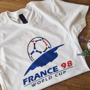 May include: White t-shirt featuring a soccer ball graphic with red, white, and blue accents, and the text "FRANCE 98 WORLD CUP" in blue and red. The shirt is a crew neck style.