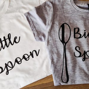 May include: Two matching t-shirts, one white and one grey, with black text and spoon graphics. The white shirt says "Little Spoon" and the grey shirt says "Big Spoon".