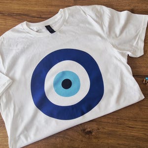 以下が含まれることがあります： 大きな青と白の目のデザインが特徴の白いTシャツ。青いタツノオトシゴ型のオーナメントと、目のシンボルが付いた小さなビーズチャームも付いています。シャツは柔らかい素材で作られています。