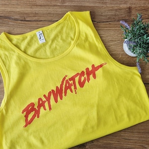 May include: A bright yellow vest top featuring the word "BAYWATCH" in red, bold lettering. The top has a round neck and is sleeveless. A small potted plant with purple flowers is placed on a wooden surface nearby.