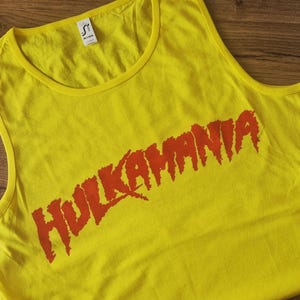 Puede incluir: Camiseta sin mangas amarilla con el texto naranja "HULKAMANIA". La camiseta sin mangas tiene cuello redondo y está hecha de un tejido suave. Una etiqueta es visible cerca del cuello. Para uso casual o deportivo.