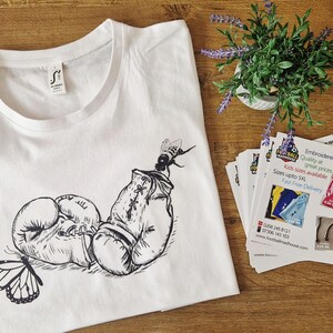 以下が含まれることがあります： ボクシンググローブ、蝶、蜂の白黒イラストが描かれた白いTシャツ。木製の表面に置かれ、小さな植物と販促カードが添えられています。
