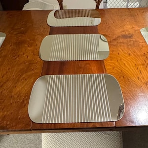 May include: A large, polished wooden dining table with a rich, warm brown finish. The table is set with five silver-coloured, rectangular placemats with vertical ribbed detailing. The table's surface has a natural wood grain pattern.
