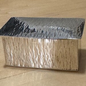 May include: A rectangular silver box with a textured surface and a hinged, angled lid. The box has a hammered appearance, giving it a rustic yet elegant look. The lid is a darker shade of silver than the body of the box.