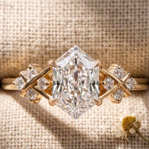 May include: A gold ring featuring a large, central hexagonal-cut clear gemstone. The band displays a crisscross design on both sides, accented with six smaller round-cut clear stones. The ring is positioned horizontally against a textured fabric.