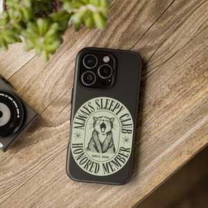 Könnte beinhalten: Schwarze Handyhülle mit einem runden Aufkleber mit der Aufschrift "ALWAYS SLEEPY CLUB HONORED MEMBER SINCE 2018". Der Aufkleber zeigt eine gähnende Katzenillustration in Hellgrün. Die Handyhülle liegt auf einer Holzoberfläche.