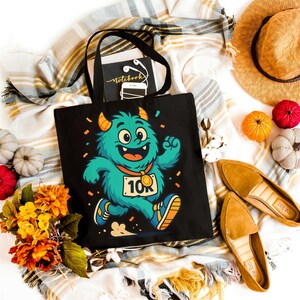 Puede incluir: Bolso tote negro con un monstruo de dibujos animados corriendo una carrera de 10 km. La imagen incluye decoraciones otoñales como calabazas, flores, un sombrero y zapatos. Un cuaderno y auriculares están dentro del bolso.