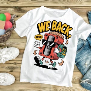 Puede incluir: Camiseta blanca con un dibujo animado de sacapuntas con gafas de sol y una mochila. El texto "WE BACK" está encima del dibujo, y "BRUH" está en un bocadillo. La camiseta está sobre una superficie de madera.