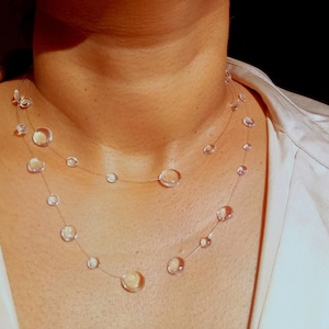 Può includere: Una delicata collana con perline rotonde trasparenti di varie dimensioni infilate su un filo quasi invisibile. La collana è a strati, con le perline che poggiano sul petto. Lo sfondo è scuro.