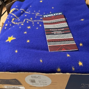 May include: A blue Pendleton blanket featuring a yellow star pattern and a black border. A product tag rests on top, reading: PENDLETON THREAD OF LIFE LEGENDARY NATIVE AMERICAN BLANKETS. The cardboard box label reads: KEEP MY FIRES BURNING, SIZE 64X80, BLUE.