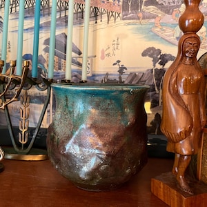 May include: A handcrafted ceramic vase with a textured surface, featuring a blend of brown and teal colors. The vase has a unique, organic shape and is displayed on a wooden surface. Decorative candles and a menorah are in the background.