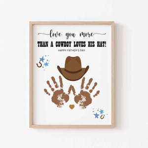 以下が含まれることがあります： 「love you more than a cowboy loves his hat!」と「Happy Father's Day」のテキストが入った額入りのプリント。茶色のカウボーイハット、手形、蹄鉄、星が茶色と青色で描かれています。