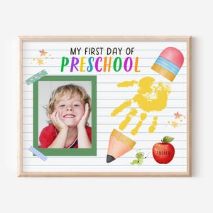 Puede incluir: Impresión enmarcada con el texto "MY FIRST DAY OF PRESCHOOL". Presenta una foto de un niño, una huella de mano amarilla, un lápiz, una manzana y elementos decorativos. La manzana tiene el nombre "Andrew".