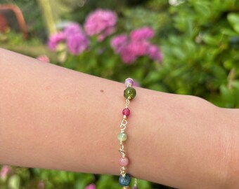 May include: A delicate bracelet featuring a gold chain adorned with a variety of colorful gemstone beads. The beads include shades of green, pink, red, and blue. The bracelet is shown on a person's arm, with a blurred floral background.