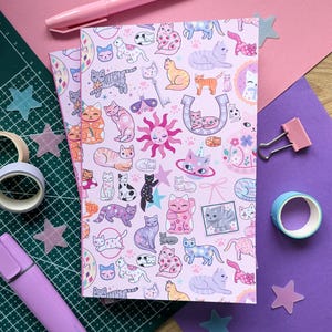 May include: A pink notebook with a playful cat design, showcasing diverse cat illustrations in various poses and colours. The pattern includes stars, keys, and a sun face. Stationery items such as tape, a marker, and star-shaped stickers are also visible.