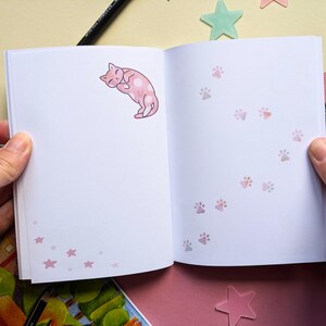 May include: An open notebook with a pink cat illustration on the left page and a trail of pink paw prints on the right page. The notebook has a white background and pink accents.