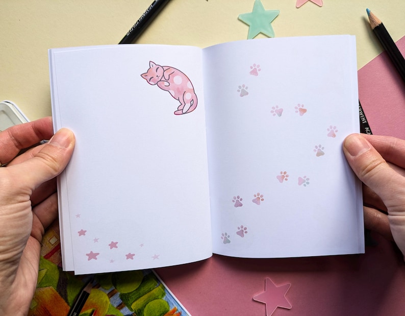 May include: An open notebook with a pink cat illustration on the left page and a trail of pink paw prints on the right page. The notebook has a white background and pink accents.