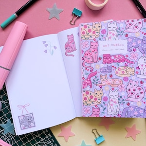 May include: An open notebook with a lavender cover featuring a pattern of illustrated cats, flowers, and strawberries. The text "Cat Cuties Illustrated Notebook" is visible. The open pages have cat and flower illustrations. Pink and teal markers and tape surround the notebook.