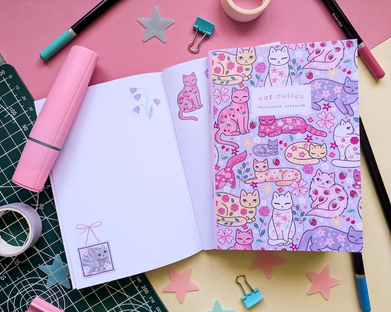 May include: An open notebook with a lavender cover featuring a pattern of illustrated cats, flowers, and strawberries. The text "Cat Cuties Illustrated Notebook" is visible. The open pages have cat and flower illustrations. Pink and teal markers and tape surround the notebook.