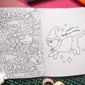 K&ouml;nnte beinhalten: Ein Malbuch, das auf zwei Seiten ge&ouml;ffnet ist. Eine Seite zeigt ein Blumendesign mit Katzen, die andere eine Katze mit Hexenhut und Umhang mit dem Text "i put a spell on meow".