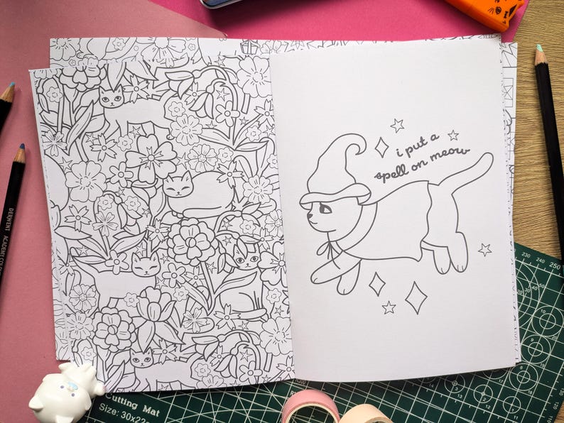 K&ouml;nnte beinhalten: Ein Malbuch, das auf zwei Seiten ge&ouml;ffnet ist. Eine Seite zeigt ein Blumendesign mit Katzen, die andere eine Katze mit Hexenhut und Umhang mit dem Text "i put a spell on meow".
