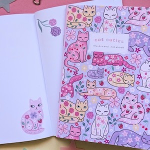 May include: An open notebook with a floral cat design. The cover reads "cat cuties illustrated notebook." The cats are decorated with flowers and strawberries on a lavender background. The left page has a cat sticker.