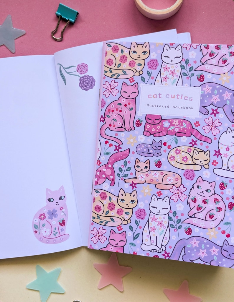 May include: An open notebook with a floral cat design. The cover reads "cat cuties illustrated notebook." The cats are decorated with flowers and strawberries on a lavender background. The left page has a cat sticker.