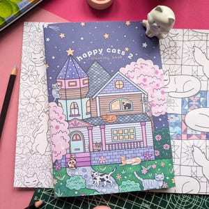 K&ouml;nnte beinhalten: Ein "Happy Cats 2" Malbuch mit einer skurrilen Hausillustration, umgeben von Katzen, Sternen und Blumen. Enth&auml;lt einen Bleistift, Klebeband und eine Katzenfigur. Die Schneidematte misst 30x21 cm.