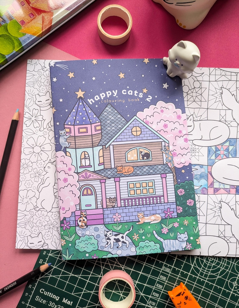 K&ouml;nnte beinhalten: Ein "Happy Cats 2" Malbuch mit einer skurrilen Hausillustration, umgeben von Katzen, Sternen und Blumen. Enth&auml;lt einen Bleistift, Klebeband und eine Katzenfigur. Die Schneidematte misst 30x21 cm.