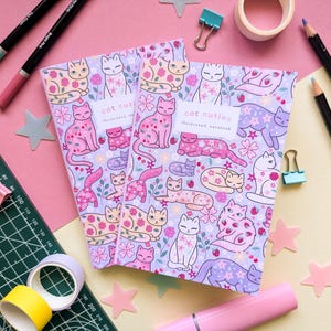May include: Two illustrated notebooks with a cat-themed design. The notebooks feature a light purple background with various cat illustrations in pink, white, and lavender, along with floral accents. The text "cat cuties" is visible on the cover.