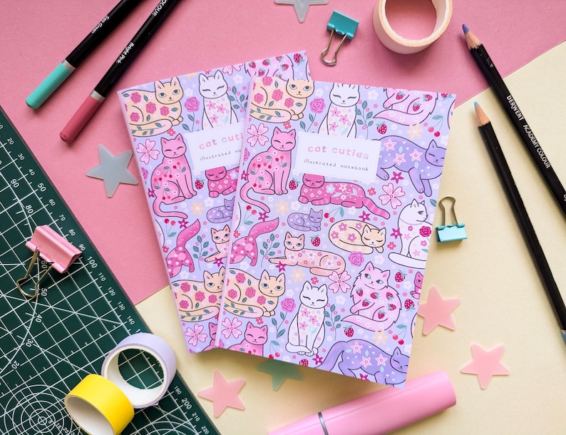 May include: Two illustrated notebooks with a cat-themed design. The notebooks feature a light purple background with various cat illustrations in pink, white, and lavender, along with floral accents. The text "cat cuties" is visible on the cover.