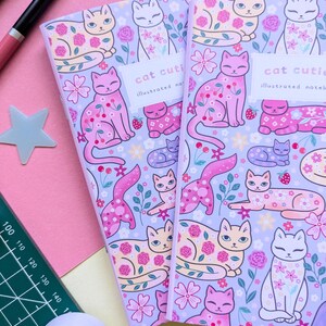 May include: Two notebooks with a lavender background and a repeating pattern of cartoon cats, flowers, and strawberries. The notebooks have the text "cat cutie" and "illustrated notebook" on the cover. Other stationery items, including pens, tape, and paperclips, are scattered around the notebooks.